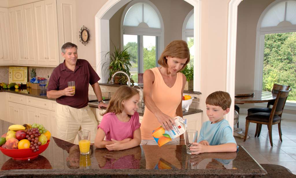 Mum in kitchen pours orange juice for son and daughter while dad watches in background