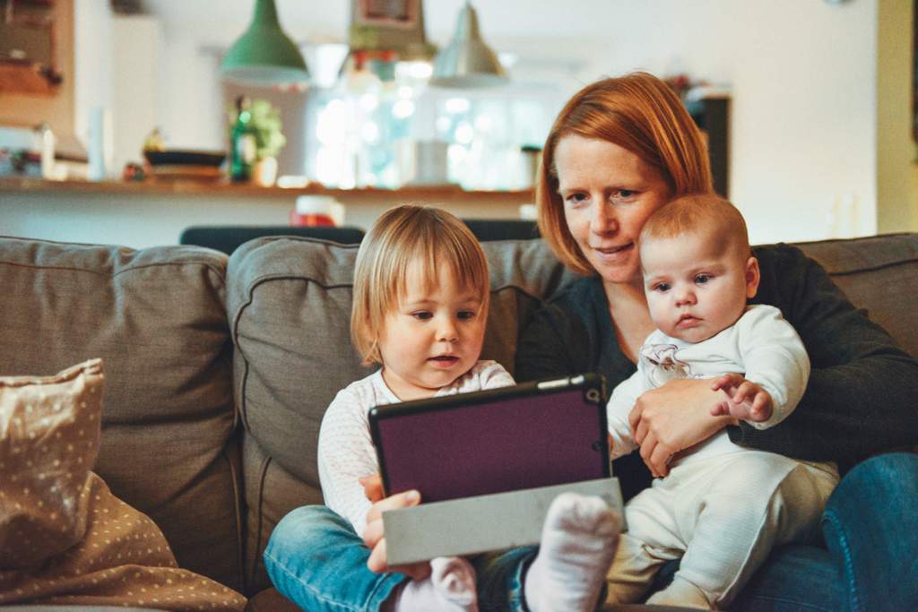 Mum sitting on sofa with toddler and baby. Toddler is holding an iPad that they're all looking at.