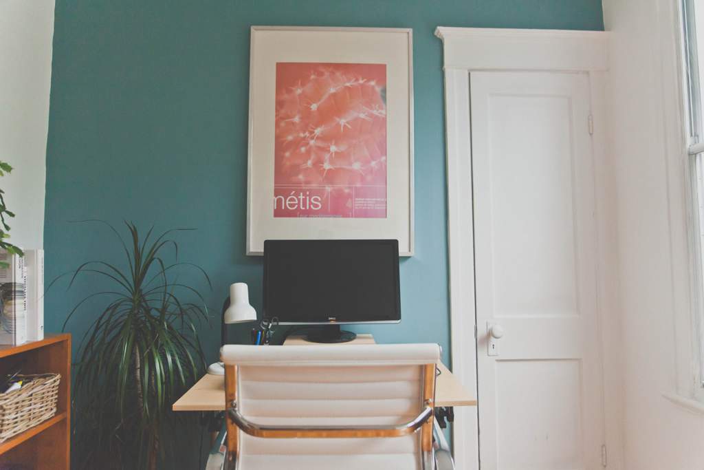 A small table and white chair facing a red picture on a blue/green wall with a white door. There is a monitor and lamp on the desk.