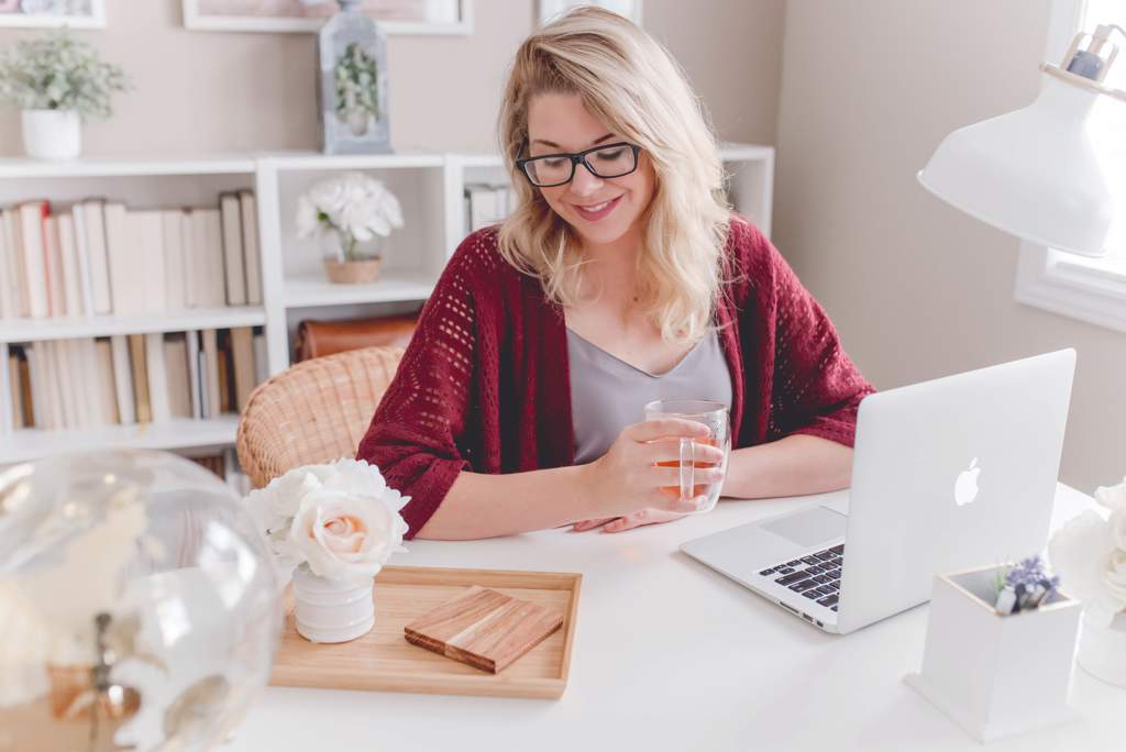 Blonde woman wearing red cardigan sitting at desk in home office with laptop open in front of her.