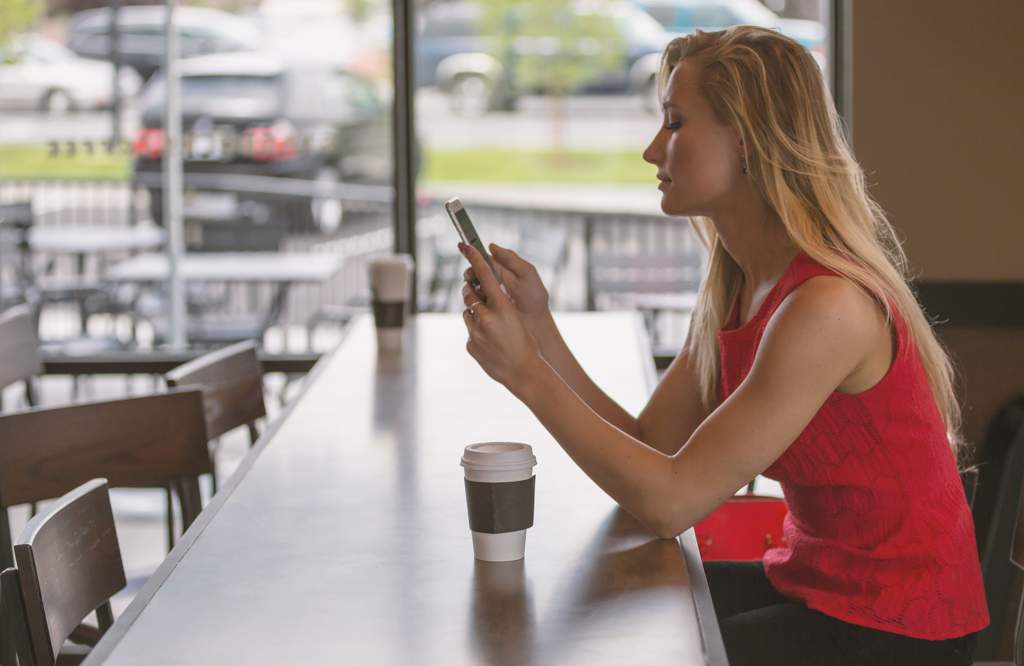 Woman with long blonde hair wearing a red sleeveless top and using a cellphone while sitting in a coffee shop with a cup of coffee on the table in front of her.