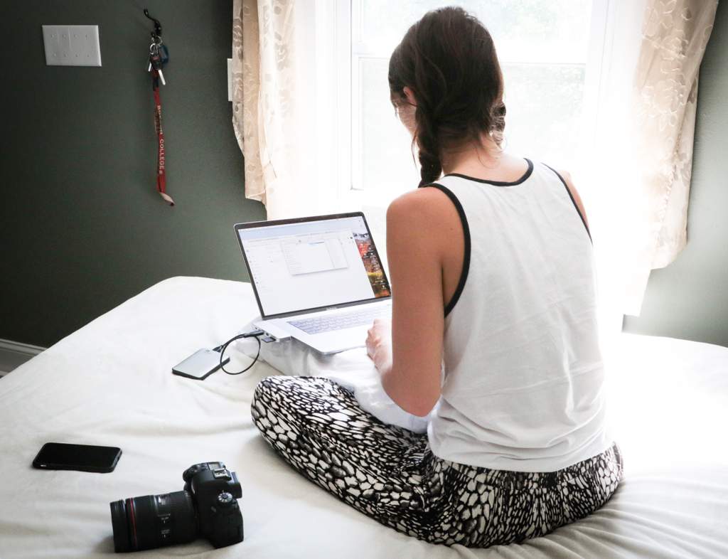 Woman sitting on a bed in front of a window with a laptop in front of her and a camera and cellphone on the bed.