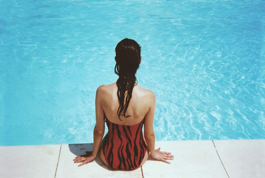Back view of a young woman with long wet hair, wearing a red and black swimming costume, sitting on the edge of a swimming pool on a sunny day.