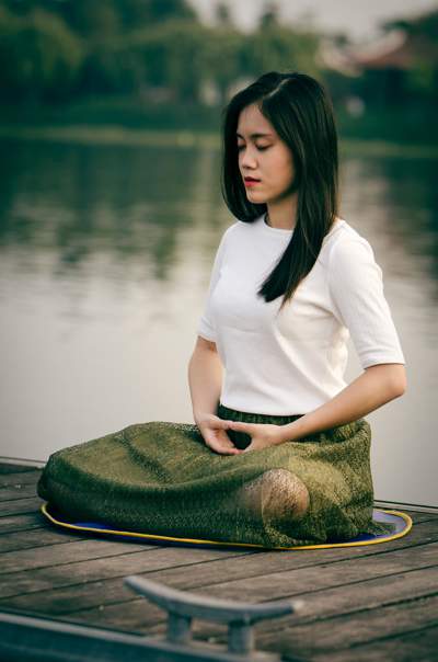Woman in a white t-shirt and green skirt sitting cross legged with her eyes closed, meditating, on a dock in front of some water.