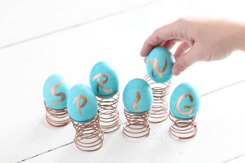 6 painted blue eggs each with a painted gold letter. Together they spell the word Spring, and each egg is sitting in a coiled copper wire egg-cup in the shape of a spring.