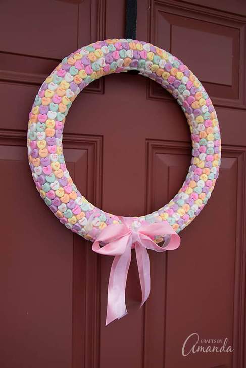Circular wreath made of candy conversation hearts with a pink ribbon bow hanging on a dark red door.