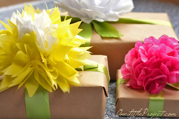 Three parcels wrapped in brown paper and tied with ribbon. Each has a large paper flower on the top, one white, one yellow with a white center and one pink.
