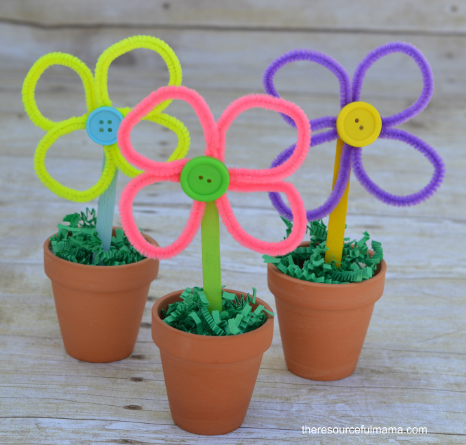 Three flowers made from pipe-cleaners in clay pots. Each flower has a craft-stick stem and a button at the centre, and the pots have green paper grass on top.
